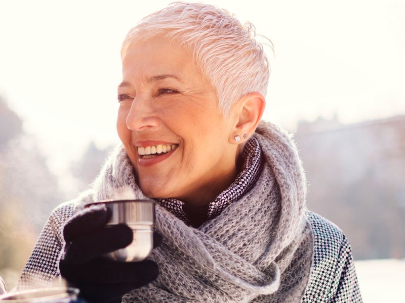 Life Extension vital mature woman with short gray hair, smiling with a cup in her hand outside on a cold sunny day